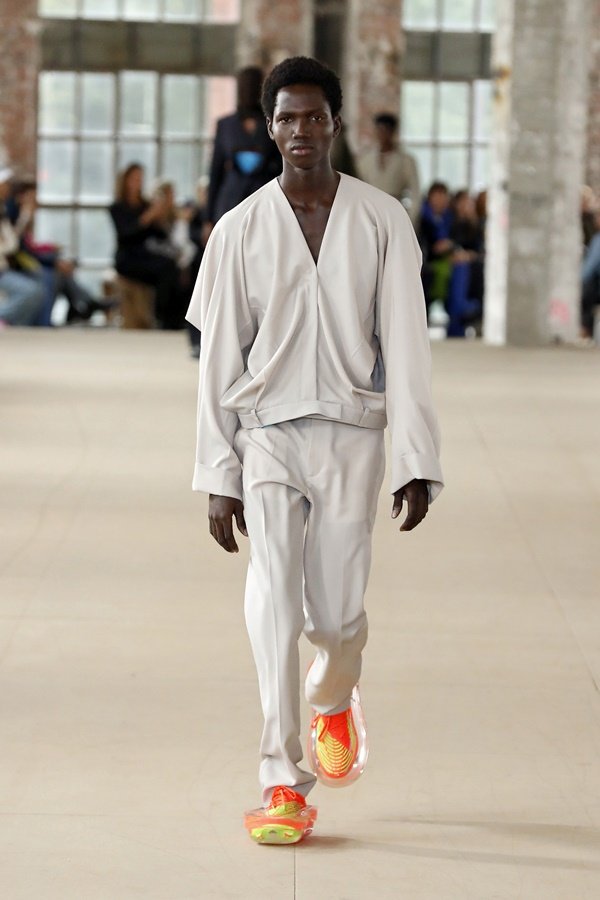 Um modelo caminha na passarela durante o desfile de moda da Botter, coleção de Primavera/Verão 2023, como parte da Paris Fashion Week em setembro de 2022. O modelo é um homem negro e jovem, de cabelo crespo curto, e usa um conjunto de casaco e calça off white e um tênis laranja