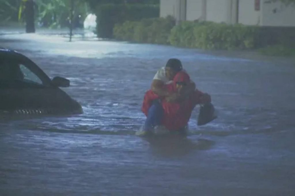 Imagem colorida de um homem carregando uma mulher nas costas em uma inundação-Metrópoles