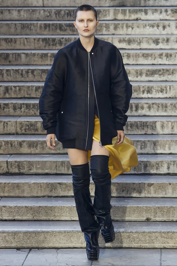 Uma modelo caminha na passarela durante o desfile de moda da marca Rick Owens, coleção de Primavera/Verão 2023, como parte da Paris Fashion Week em setembro de 2022 em Paris, França. A modelo é uma mulher branca e jovem, de cabelo preto raspado, que veste uma saia amarela e um casaco preto.