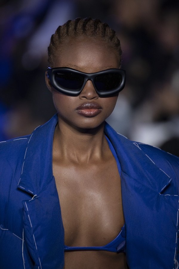 Uma modelo caminha na passarela durante o desfile de moda da marca Off-White, coleção de Primavera/Verão 2023, como parte da Paris Fashion Week em setembro de 2022 em Paris, França. A modelo é uma mulher negra e jovem, de cabelos traçados, e usa um top preto, blazer azul jeans e um óculos escuro preto