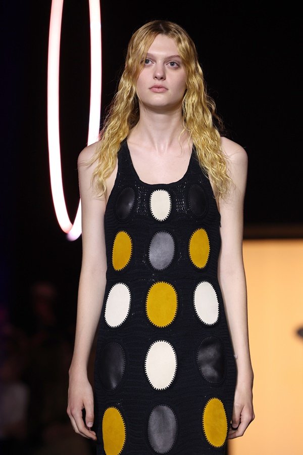 Uma modelo caminha na passarela durante o desfile de moda da marca Chloé, coleção de Primavera/Verão 2023, como parte da Paris Fashion Week em setembro de 2022 em Paris, França. A modelo é uma mulher branca e jovem, de cabelos lisos loiros, e usa um vestido preto com bolas brancas e amarelas.