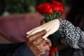 Mão de homem negro colocando anel em dedo de mulher branca e buquê de rosas vermelhas ao fundo