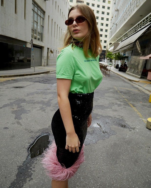 Mulher branca e jovem, de cabelo loiro liso, posando para foto nas ruas de São Paulo. Ela usa uma camiseta verde, óculos escuros e uma saia preta de paetê com plumas rosas na barra