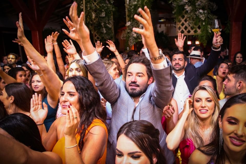 A foto colorida mostra o cantor Fernando Zor usando um terno cinza com uma blusa social branca por dentro. Ele está pulando com as mãos e braços para cima, tentando pegar o buquê do casamento de João Bosco. Outras pessoas ao redor fazem o mesmo
