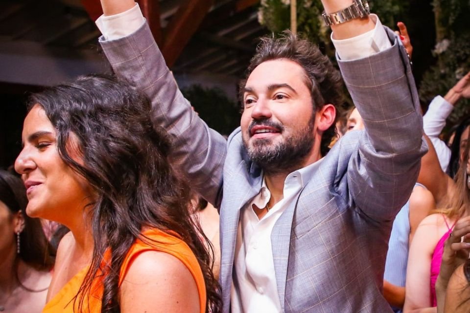 A foto colorida mostra o cantor Fernando Zor usando um terno cinza com uma blusa social branca por dentro. Ele está pulando com as mãos e braços para cima, tentando pegar o buquê do casamento de João Bosco. Outras pessoas ao redor fazem o mesmo
