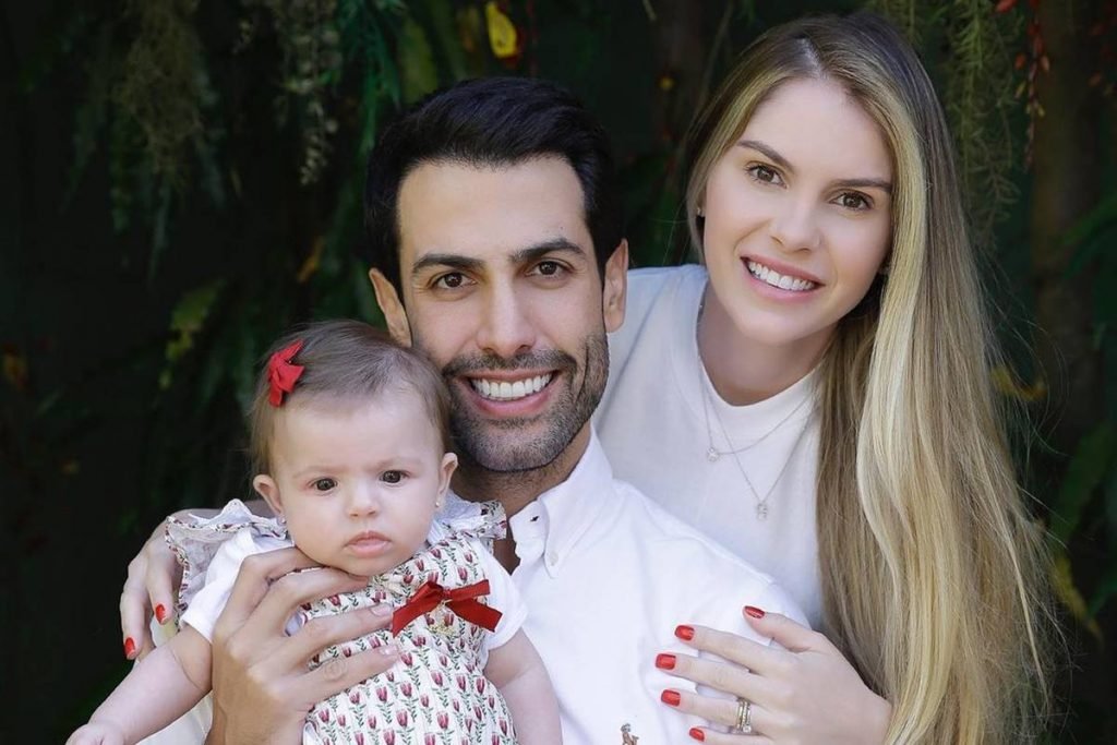 Mulher loura sorri atrás de homem branco de cabelos pretos e barba rala segurando uma bebe com vestido branco com estampas vermelhas e laço na cabeça