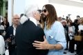 O estilista Karl Lagerfeld, um homem branco, idoso e de cabelos brancos e lisos comprimentando o ator Jared Leto, um homem branco de cabelo liso longo castanho. A foto é de 2014 em um desfile da marca Chanel.