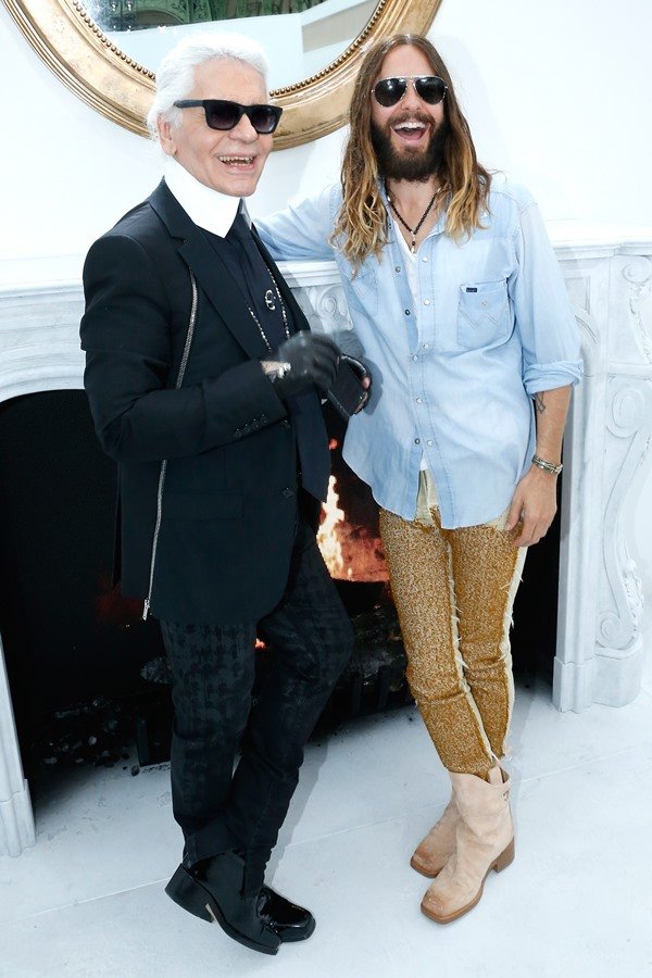 O estilista Karl Lagerfeld, um homem branco, idoso e de cabelos brancos e lisos comprimentando o ator Jared Leto, um homem branco de cabelo liso longo castanho. A foto é de 2014 em um desfile da marca Chanel.