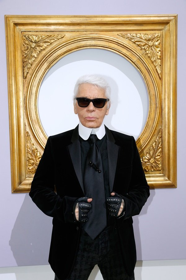 O estilista Karl Lagerfeld, um homem idoso, branco e de cabelos brancos lisos, posando para foto em frente a uma moldura dourada. Ele usa um terno preto e luvas pretas de couro. A foto é de um desfile da Chanel de 2014.