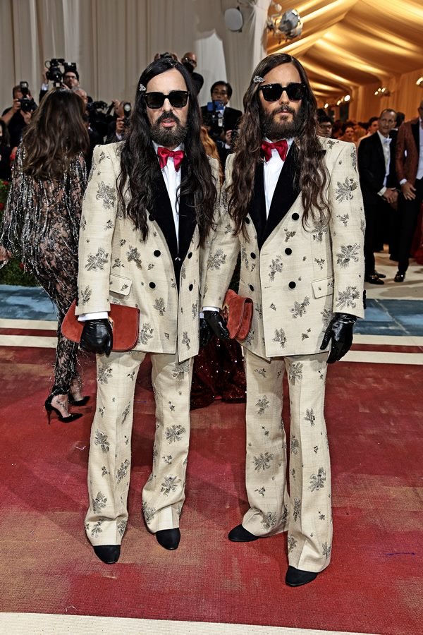 O estilista Alessandro Michele e o ator Jared Leto, vestidos iguais, no Met Gala de 2022 em Nova York, nos Estados. Eles são brancos, usam barba e tem cabelos castanhos longos e vestem um terno branco com estampa prata.