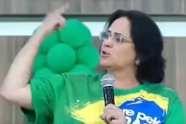 Fotografia colorida de mulher com camiseta do Brasil discursando com microfone na mão esquerda