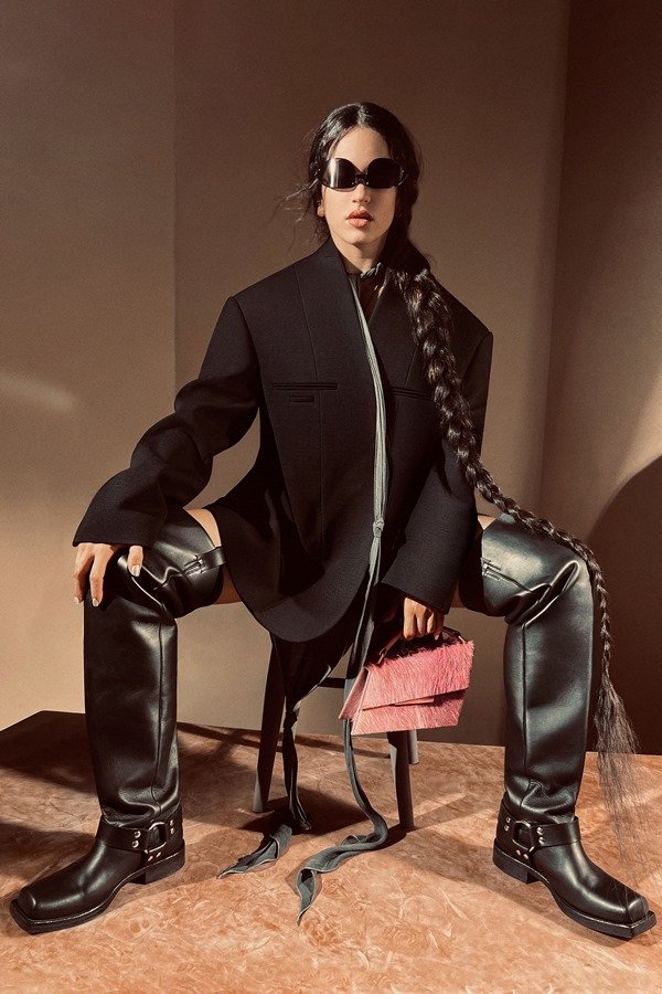 A cantora Rosalía, uma mulher jovem, branca, de cabelo ondulado longo, posando para a campanha da marca Acne Studios. Ela está em um estúdio com paredes marrons a e usa um blazer preto grande, óculos escuros, bota grande de couro e uma bolsa rosa.