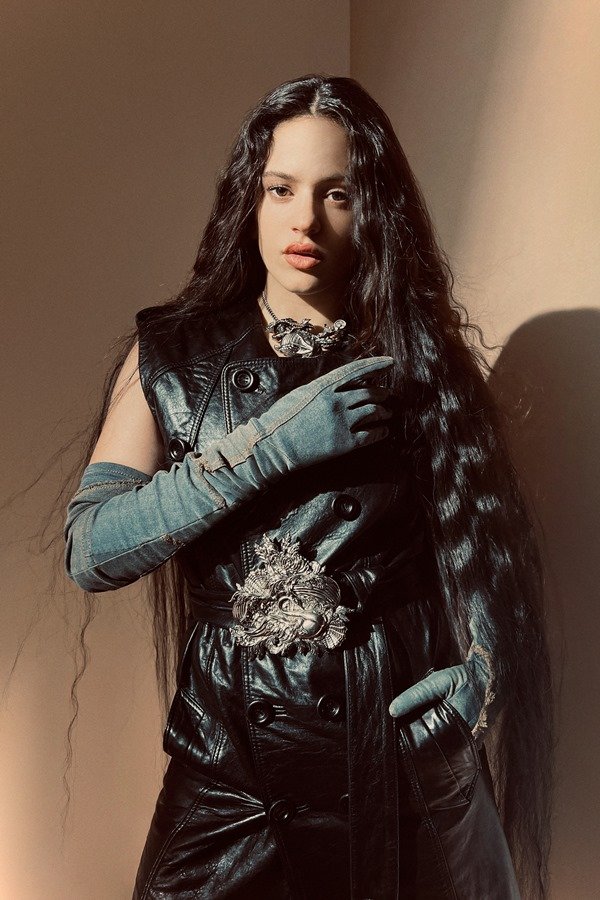 A cantora Rosalía, uma mulher jovem, branca, de cabelo ondulado longo, posando para a campanha da marca Acne Studios. Ela está em um estúdio com paredes marrons a e usa um casaco preto de couro e um cinto com fivela prata.
