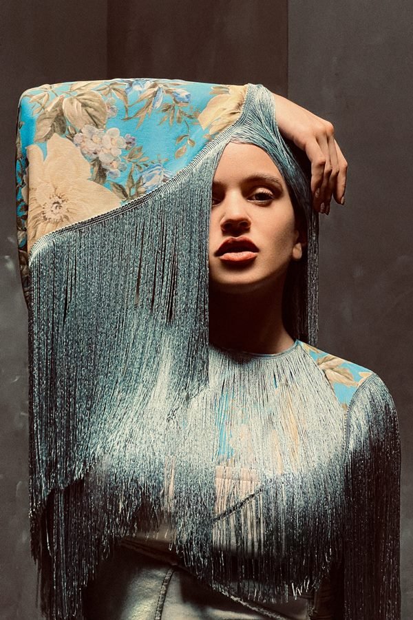  A cantora Rosalía, uma mulher jovem, branca, de cabelo ondulado longo, posando para a campanha da marca Acne Studios. Ela está em um estúdio com paredes marrons a e usa um vestido com estampa de flores azuis e amarelas com detalhes de estrasse.