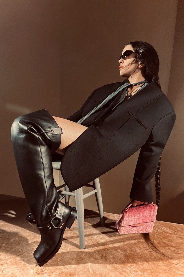 A cantora Rosalía, uma mulher jovem, branca, de cabelo ondulado longo, posando para a campanha da marca Acne Studios. Ela está em um estúdio com paredes marrons a e usa um blazer preto grande, óculos escuros, bota grande de couro e uma bolsa rosa.
