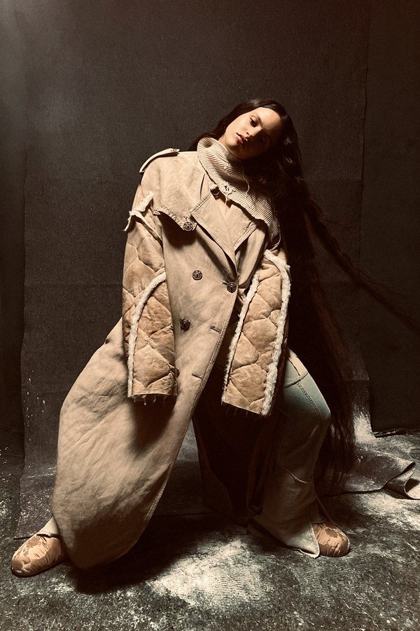 A cantora Rosalía, uma mulher jovem, branca, de cabelo ondulado longo, posando para a campanha da marca Acne Studios. Ela está em um estúdio com paredes marrons a e usa um casaco marrom acolchoado com detalhes de pelo branco.
