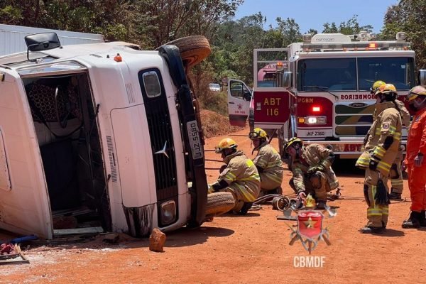 Caminhão branco tombado em terra e bombeiros ao lado