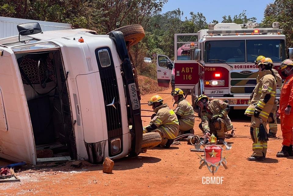 Caminhão branco tombado em terra e bombeiros ao lado
