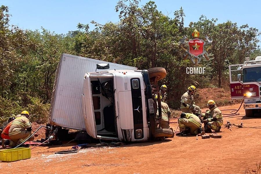 Caminhão branco tombado em terra e bombeiros ao lado