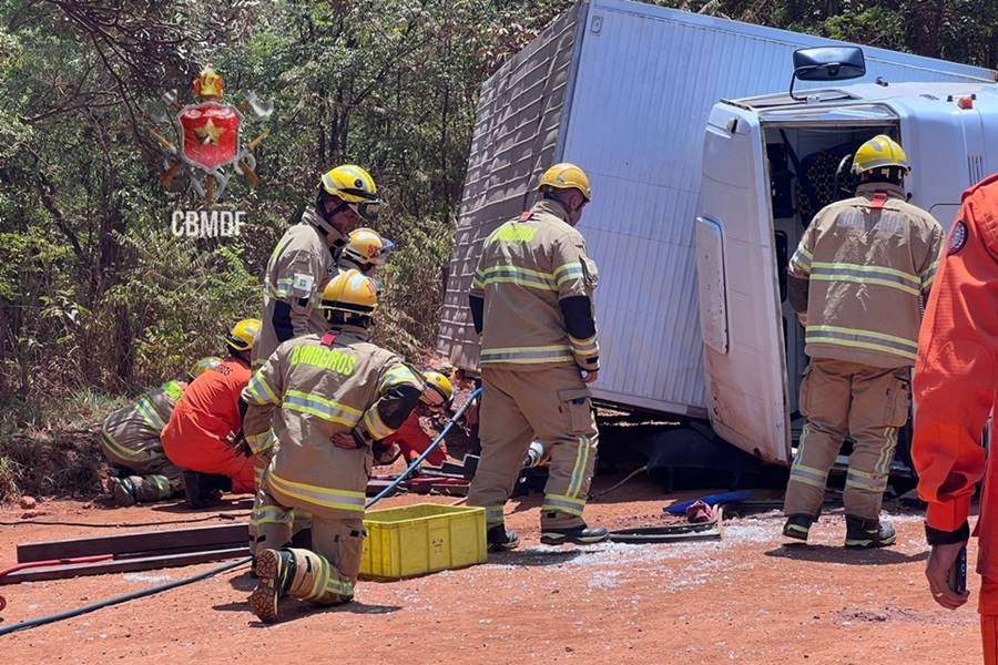 Caminhão branco tombado em terra e bombeiros ao lado