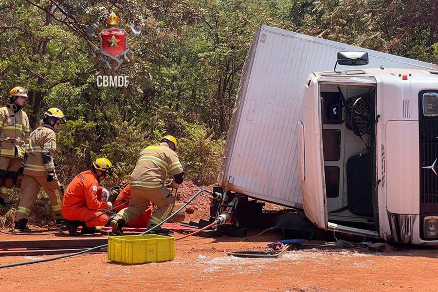 Caminhão branco tombado em terra e bombeiros ao lado