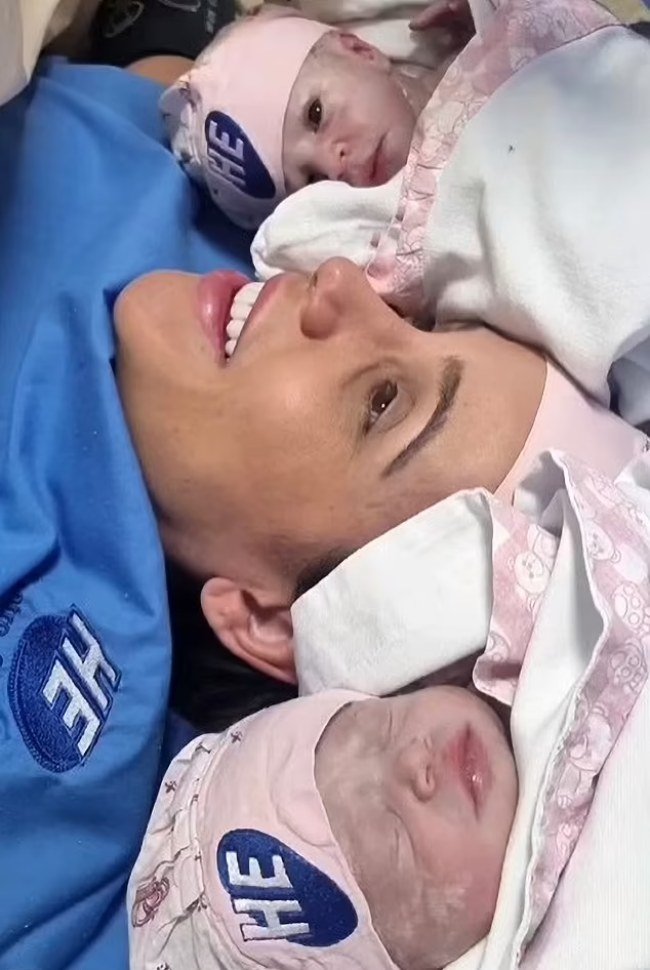 fotografia colorida de uma mãe entre as duas filhas gêmeas após nascerem. A ,ãe está com as roupas do hospital e as bebês estão com touquinhas rosa