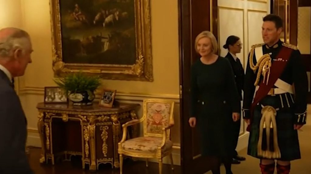 Foto colorida de mulher branca e loira entrando em uma sala com quadros. Ao lado dela, um homem com trajes militares - Metrópoles