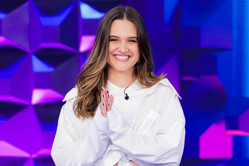 Juliana Paiva participa do Jogo das 3 Pistas no Programa Silvio Santos. Na foto ela sorri, usando blusa branca e bate palmas frente a fundo roxo e azul - Metrópoles