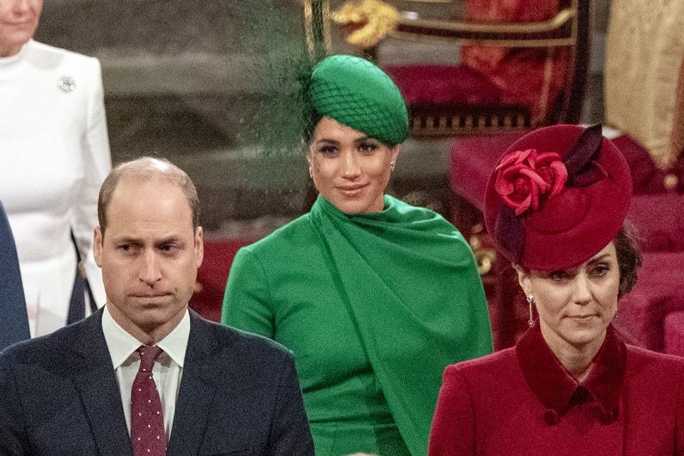 Foto colorida de homem branco e careca ao lado de mulher branca com vestido e chapéu vermelho. Ao fundo, mulher parda com roupas e chapéu verde - Metrópoles