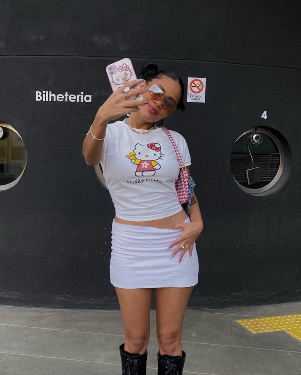Mulher jovem e negra, de cabelo amarrado em coque, posando para foto nas ruas de São Paulo. Ela usa uma camiseta branca com estampa da gata Hello Kitty, saia branca curta, bolsa rosa e óculos com lente furta-cor.