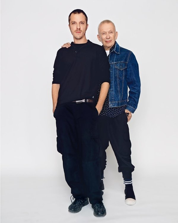 Os estilistas Jean-Paul Gaultier e Glenn Martens posando para foto em um fundo branco. Ambos são brancos e usam calças jeans com camiseta, mas Gleen é jovem e Jean-Paul já é idoso - Metrópoles