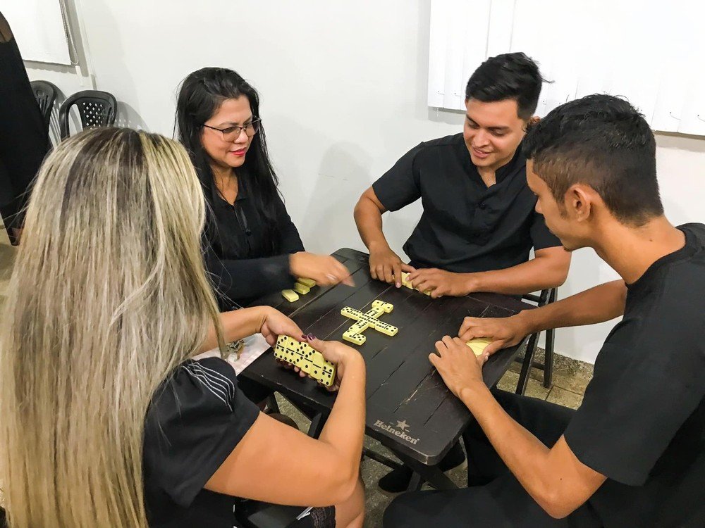 foto colorida de pessoas de preto sentadas ao redor da mesa jogando domin[o