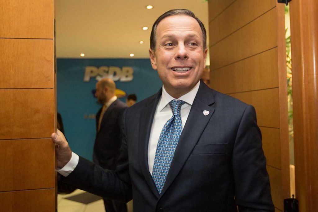O ex-governador de São Paulo e empresário, João Dória, sorri na porta de um comitê do PSDB. É possível ver outras pessoas ao fundo e a logo do partido em parede azul - Metrópoles