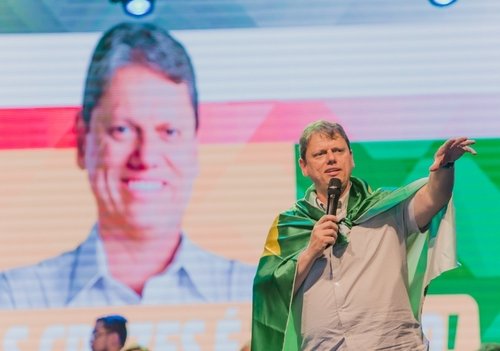O candidato ao governo de São Paulo, Tarcísio de Freitas, fala em ato de campanha com microfone na mão. Ele usa bandeira do Brasil amarrada no pescoço com capa diante de um telão com sua foto atrás - Metrópoles