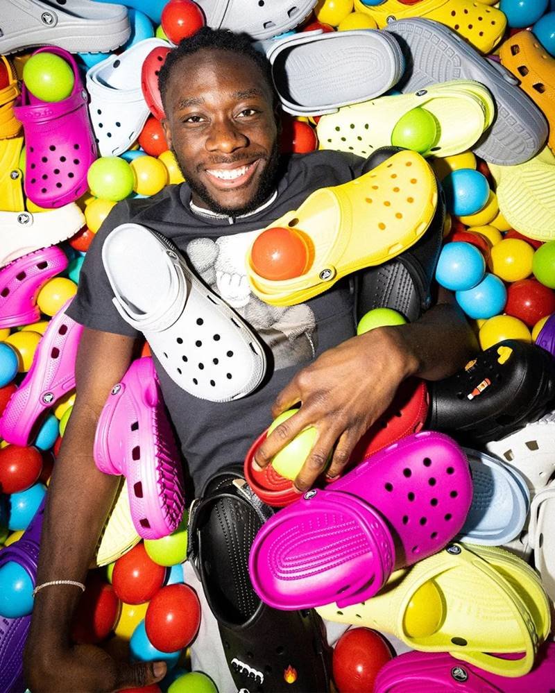 Homem negro e jovem posando para foto em campanha oficial da marca de sapatos Crocs. Ele está em uma piscina de bolinha com vários sapatos da empresa coloridos - Metrópoles