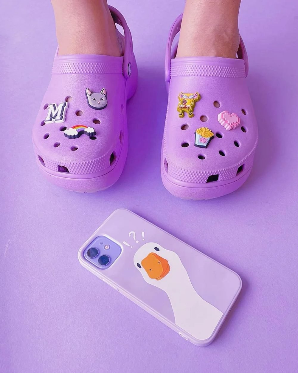 Sapatos da marca Crocs na cor lilás, em um fundo de mesmo tom, com um celular iPhone 13 com capinha na mesma cor - Metrópoles