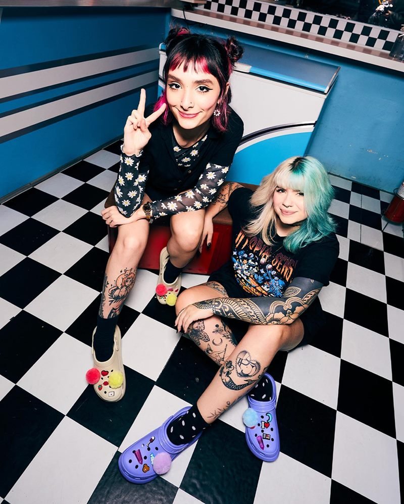 Duas mulheres jovens e de cabelo liso colorido posando para foto em uma campanha da marca Crocs. Elas usam roupas pretas, possuem tatuagens e usam meias, também pretas, com os sapatos da marca coloridos: um rosa e um lilás - Metrópoles