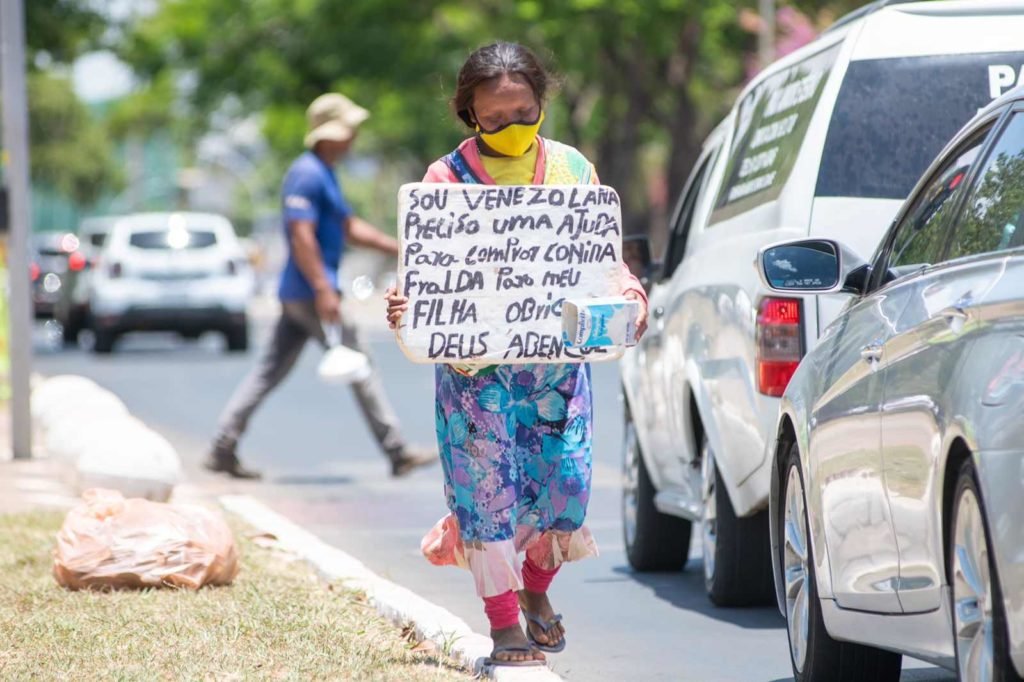 Refugiada venezuelana Herminia Zapata percorre semáforos do DF para conseguir dinheiro com caixinha e placa. Ela usa vestido florido e máscara amarela - Metrópoles