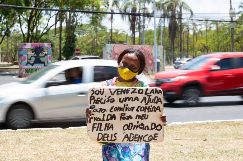 Refugiada venezuelana Herminia Zapata percorre semáforos do DF para conseguir dinheiro com caixinha e placa. Ela usa vestido florido e máscara amarela - Metrópoles
