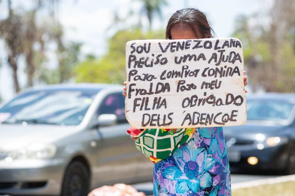 Mulher venezuelana com vestido colorido e máscara segurando placa com os dizeres: