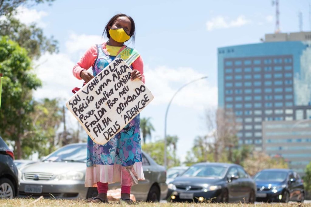 Mulher venezuelana com vestido colorido e máscara segurando placa com os dizeres: