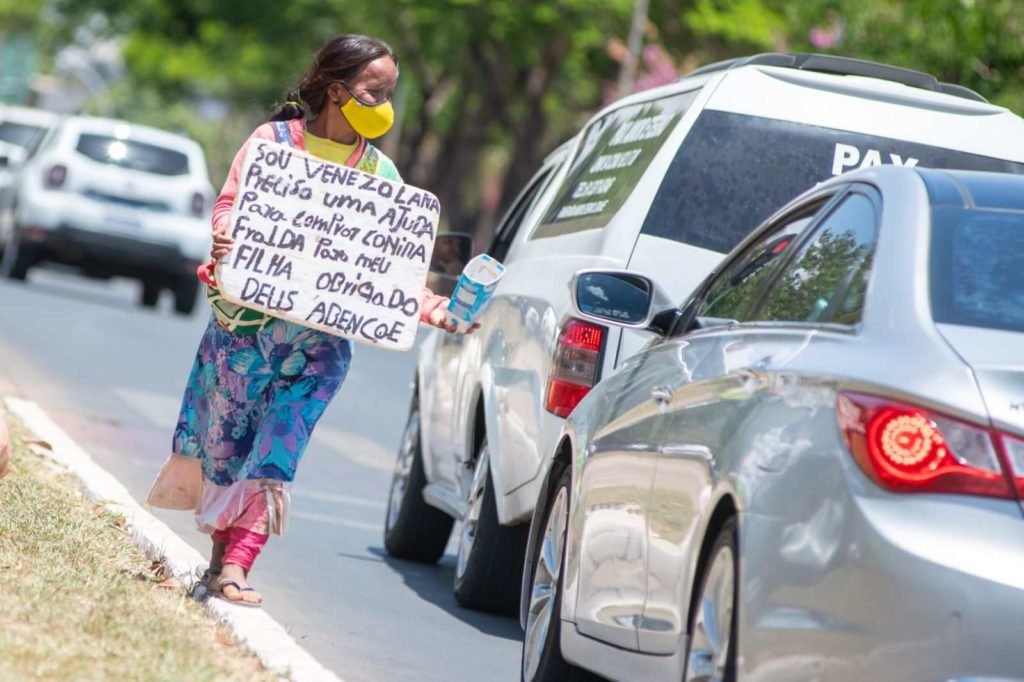 Refugiada venezuelana Herminia Zapata percorre semáforos do DF para conseguir dinheiro com caixinha e placa. Ela usa vestido florido e máscara amarela - Metrópoles