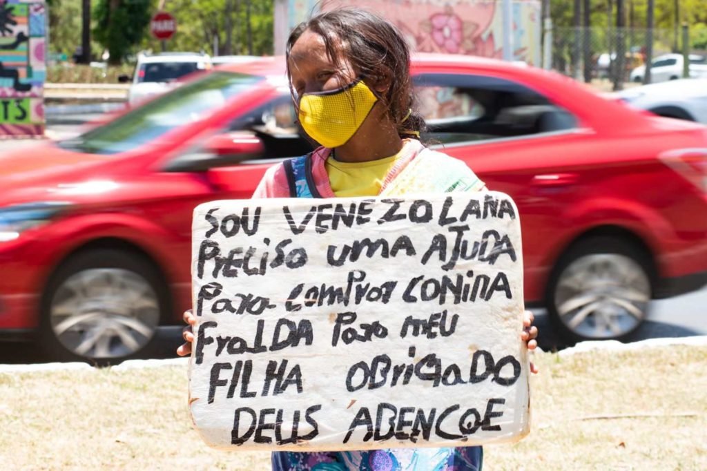 Refugiada venezuelana Herminia Zapata percorre semáforos do DF para conseguir dinheiro com caixinha e placa. Ela usa vestido florido e máscara amarela - Metrópoles