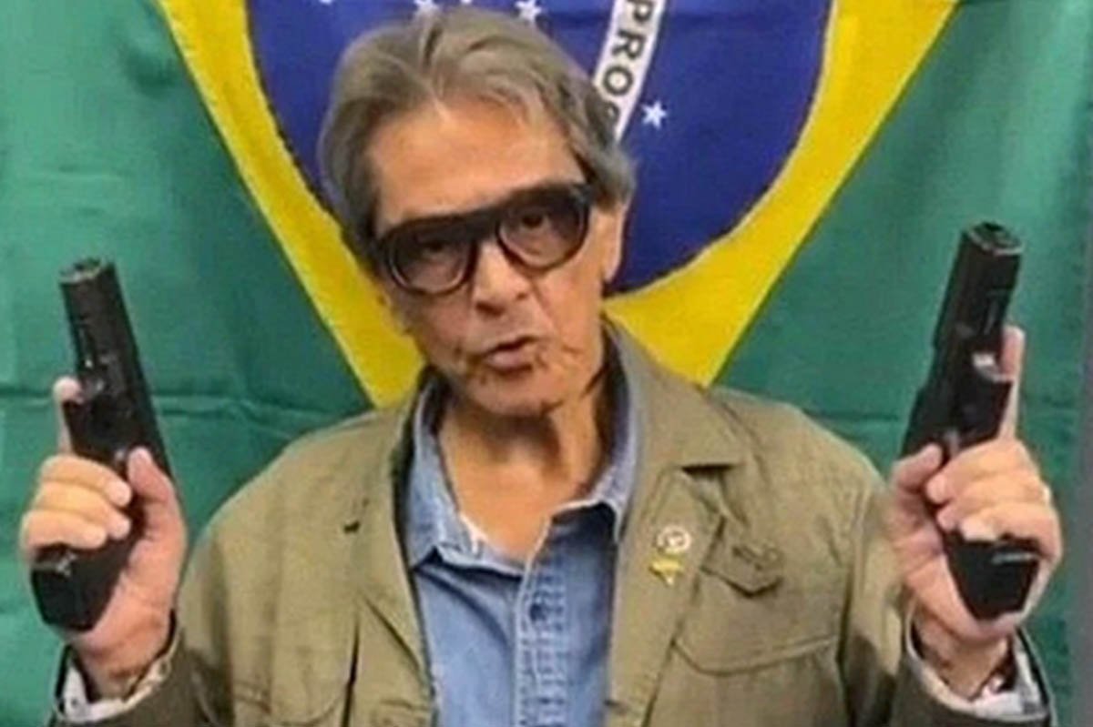 Roberto Jefferson, ex-deputado e bolsonarista, segura dois revólveres apontados para cima com a bandeira do Brasil atrás enquanto faz discurso - Metrópoles