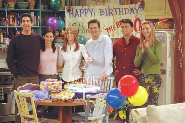 Foto colorida com o elenco principal de FRiends em frente a uma mesa com balões e cartazes