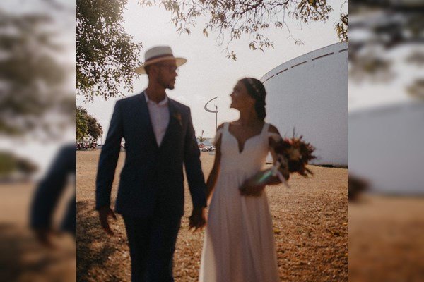 Casal de noivos. Ela com um buquê nos braços e vestido branco. Ele de blazer azul, camisa e chapéu branco - Metrópoles