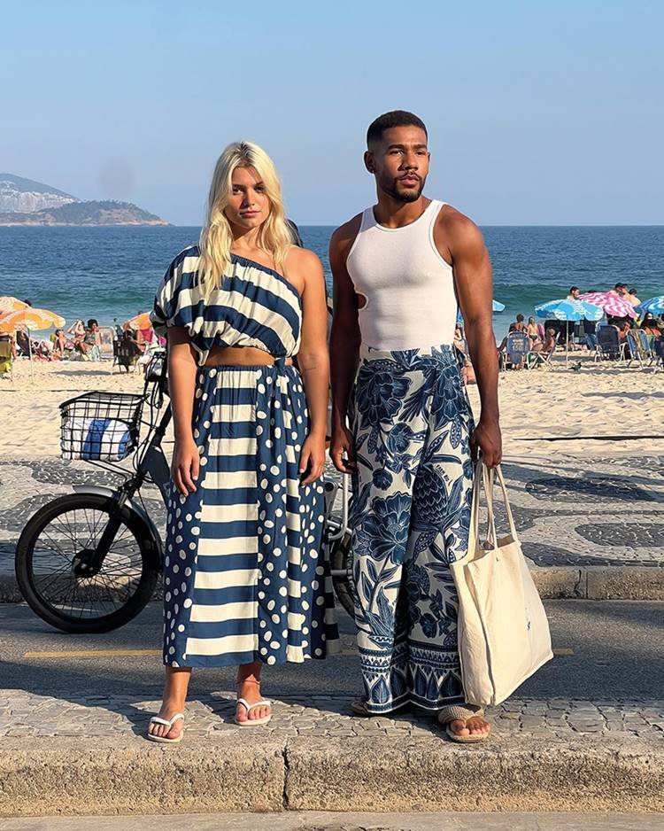 Uma mulher branca, jovem e loira, ao lado de um homem jovem, negro e com o cabelo curto, ambos no calçadão da praia do Rio de Janeiro. Eles usam roupas nas cores azul e branco da marca Farm