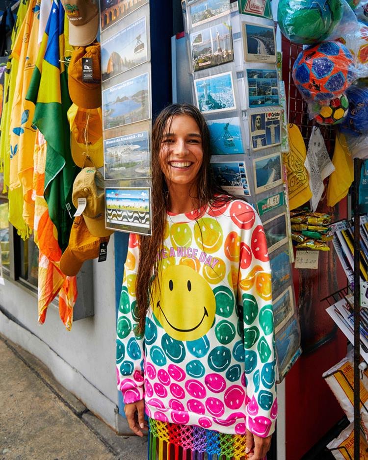 Mulher branca e jovem, de cabelos lisos longos, posando para foto em um camelô de produtos turísticos no Rio de Janeiro. Ela usa um moletom colorido da marca Farm em parceria com o personagem Smiley.