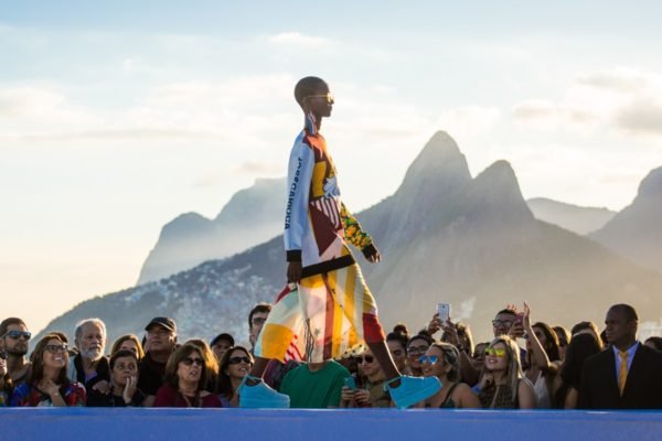 Mulher jovem, negra e de cabelo raspado cruzando a passarela com looks da Farm. A estrutura é montada em um local no Rio que tem o Morro do Pão de Açúcar ao fundo. É possível ver pessoas na plateia assistindo ao show