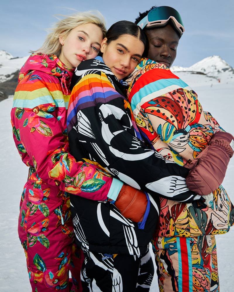 Foto de campanha da marca Farm na neve. São roupas feitas para o inverno. Na imagem é possível ver três modelos: uma branca e loira, outra morena e a terceira é negra. Usam macacões estampados.